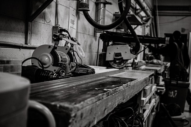 A workshop scene featuring industrial tools and machinery, including a saw on a workbench. In the background, a person, possibly a worker, is partially visible among various equipment. The setting is utilitarian and functional, with cables and metal pipes visible along the walls, contributing to a sense of busyness and productivity.