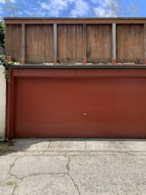 A red garage door is set against a backdrop of a wooden fence. The concrete ground in front displays visible cracks and patches of greenery. The fence is weathered, suggesting exposure to the elements, and clouds dot the blue sky above.