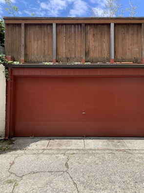 A red garage door is set against a backdrop of a wooden fence. The concrete ground in front displays visible cracks and patches of greenery. The fence is weathered, suggesting exposure to the elements, and clouds dot the blue sky above.