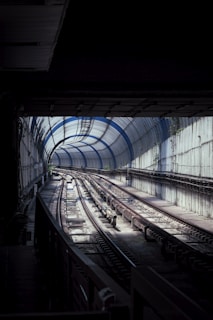 A dimly lit railway tunnel with metal tracks curving into the distance, partially illuminated by natural daylight filtering through a series of arched, transparent panels above. The tunnel is flanked by concrete walls with patches of greenery visible in some areas.