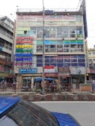 A multi-story commercial building with colorful signs in various scripts covering the facade. The building has glass windows displaying clothing inside. In front of the building, there are rickshaws, parked vehicles, and pedestrians on the street.