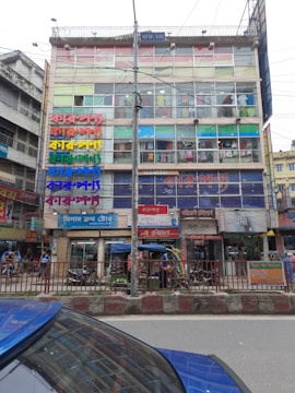 A multi-story commercial building with colorful signs in various scripts covering the facade. The building has glass windows displaying clothing inside. In front of the building, there are rickshaws, parked vehicles, and pedestrians on the street.
