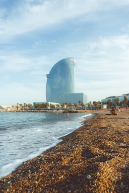 A large, modern hotel building with a glass facade is situated near a beach. The sandy shoreline stretches alongside a calm sea, with a number of people enjoying the sunny weather. Palm trees line the promenade, adding to the coastal atmosphere, and the sky is mostly clear with some light clouds.