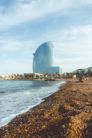 A large, modern hotel building with a glass facade is situated near a beach. The sandy shoreline stretches alongside a calm sea, with a number of people enjoying the sunny weather. Palm trees line the promenade, adding to the coastal atmosphere, and the sky is mostly clear with some light clouds.