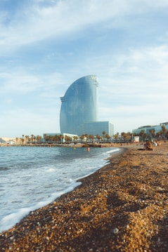 A large, modern hotel building with a glass facade is situated near a beach. The sandy shoreline stretches alongside a calm sea, with a number of people enjoying the sunny weather. Palm trees line the promenade, adding to the coastal atmosphere, and the sky is mostly clear with some light clouds.
