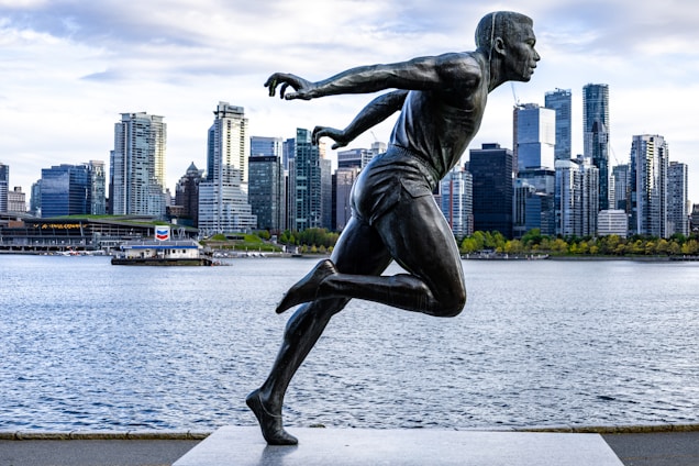 A dynamic statue of a running athlete is positioned prominently in the foreground with a backdrop of a city skyline. The cityscape includes numerous modern high-rise buildings along a waterfront, with clear skies overhead and calm water reflecting the structures.