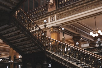 An ornate and richly decorated interior featuring a grand staircase with elaborate wrought iron railings. The architecture includes detailed moldings, columns, and intricate patterns. The lighting consists of globe-shaped lamps, adding to the vintage and luxurious atmosphere.