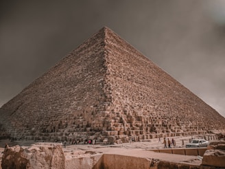 A large, ancient stone pyramid with a prominent triangular shape stretches into a cloudy sky. The structure is composed of massive stone blocks. People are visible at the base, providing a sense of scale, while vehicles are parked nearby. The lighting is subdued, contributing to a historic ambiance.