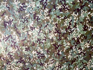 A detailed camouflage pattern consisting of irregular shapes and colors that blend into an abstract design. The colors include various shades of green, brown, and beige with dark and light accents.