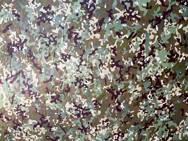 A detailed camouflage pattern consisting of irregular shapes and colors that blend into an abstract design. The colors include various shades of green, brown, and beige with dark and light accents.