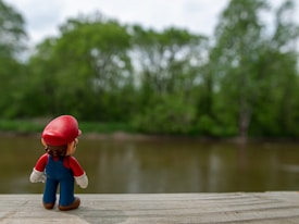 A figurine of a character wearing a red hat and blue overalls stands on a wooden surface, overlooking a calm, brown river surrounded by lush green trees.