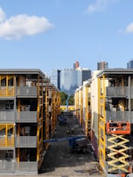 Two adjacent modern apartment buildings with yellow and gray accents. In the middle, a construction site is visible with workers and equipment, including a scissor lift and crane. In the background, a city skyline with various skyscrapers can be seen against a clear blue sky.
