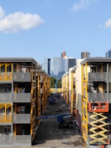 Two adjacent modern apartment buildings with yellow and gray accents. In the middle, a construction site is visible with workers and equipment, including a scissor lift and crane. In the background, a city skyline with various skyscrapers can be seen against a clear blue sky.