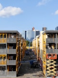 Two adjacent modern apartment buildings with yellow and gray accents. In the middle, a construction site is visible with workers and equipment, including a scissor lift and crane. In the background, a city skyline with various skyscrapers can be seen against a clear blue sky.
