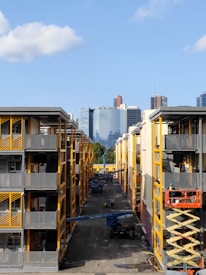 Two adjacent modern apartment buildings with yellow and gray accents. In the middle, a construction site is visible with workers and equipment, including a scissor lift and crane. In the background, a city skyline with various skyscrapers can be seen against a clear blue sky.