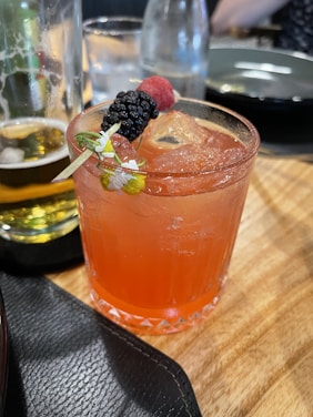 A vibrant red cocktail is garnished with a skewer of fresh berries, including a blackberry and a raspberry, and a small flower. The drink is served in a textured glass filled with ice cubes. In the background, there is a partially visible beer glass with some foam, a clear bottle of water, and a black plate on a wooden table.