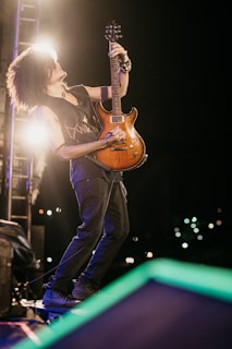 A musician with curly hair passionately plays an electric guitar on stage. The scene is set in low light, with spotlights creating a dramatic effect. The musician is wearing a sleeveless shirt and is fully engaged in the performance.