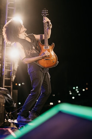 A musician with curly hair passionately plays an electric guitar on stage. The scene is set in low light, with spotlights creating a dramatic effect. The musician is wearing a sleeveless shirt and is fully engaged in the performance.