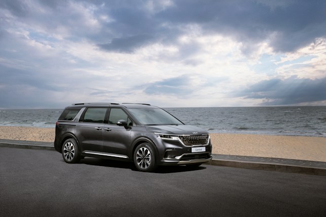 A dark-colored minivan is parked on a paved road by the beach. The ocean stretches into the horizon under a cloudy sky with some sunlight breaking through.