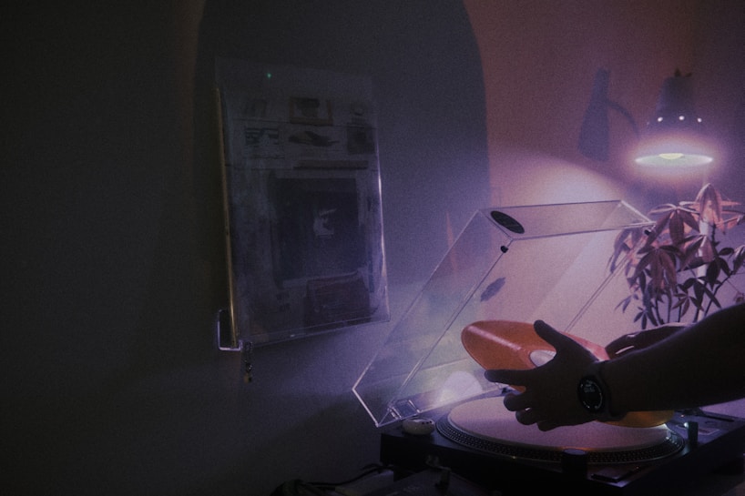 A dimly lit room with a focus on a turntable where a person is handling a translucent orange vinyl record. There is a houseplant and ambient light from a lamp casting soft shadows, creating a moody atmosphere.