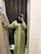 A person wearing a green embroidered gown stands in front of a mirror. The gown features intricate gold patterns and embellishments. The individual has a blue hijab and is capturing the image with a smartphone. A similar dress in a different color hangs on a rack beside them.