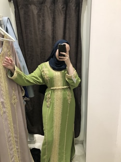 A person wearing a green embroidered gown stands in front of a mirror. The gown features intricate gold patterns and embellishments. The individual has a blue hijab and is capturing the image with a smartphone. A similar dress in a different color hangs on a rack beside them.