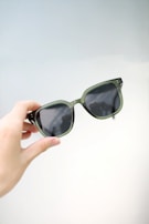 A close-up of a hand holding a contact lens with a blurred background of designer spectacle frames.