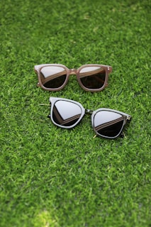Two pairs of sunglasses are placed on a lush green grass surface. One pair has a light brown frame with rectangular lenses, while the other pair features a dark frame with more rounded lenses.