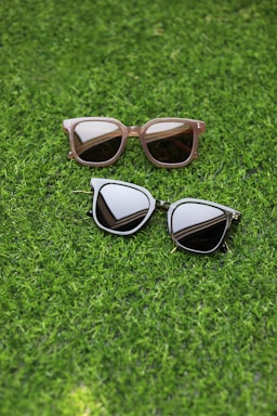 Two pairs of sunglasses are placed on a lush green grass surface. One pair has a light brown frame with rectangular lenses, while the other pair features a dark frame with more rounded lenses.