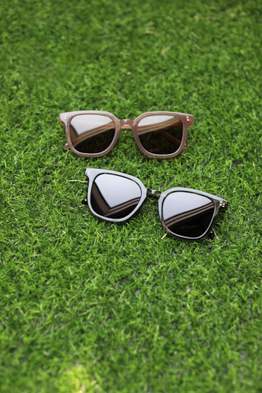 Two pairs of sunglasses are placed on a lush green grass surface. One pair has a light brown frame with rectangular lenses, while the other pair features a dark frame with more rounded lenses.