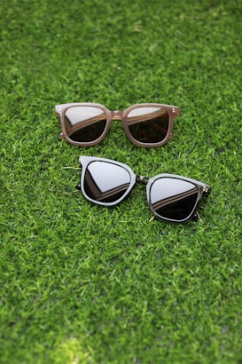 Two pairs of sunglasses are placed on a lush green grass surface. One pair has a light brown frame with rectangular lenses, while the other pair features a dark frame with more rounded lenses.