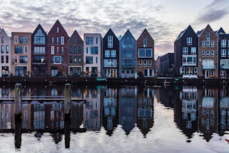 Row of modern residential buildings with distinct triangular roofs reflected in a calm body of water. The architecture includes a mix of brick and glass facades. Wooden posts are visible in the foreground, adding to the serene waterway setting beneath a cloudy sky.