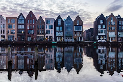 Row of modern residential buildings with distinct triangular roofs reflected in a calm body of water. The architecture includes a mix of brick and glass facades. Wooden posts are visible in the foreground, adding to the serene waterway setting beneath a cloudy sky.