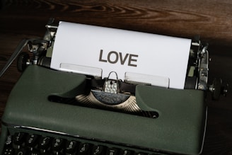 A vintage typewriter with a sheet of paper emerging from it bearing the word 'LOVE' in bold, uppercase letters. The typewriter is placed on a wooden surface, giving a nostalgic and classic appearance.