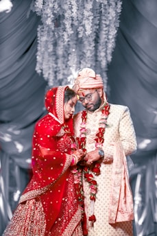 A couple stands closely together, dressed in traditional Indian wedding attire. The bride is wearing a bright red lehenga with intricate embroidery and has a veil covering her head. The groom is in a cream sherwani with a matching turban. Both are adorned with flower garlands. They appear to be in a decorative setting with draped fabric and hanging floral arrangements in the background.