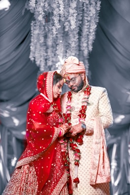 A couple stands closely together, dressed in traditional Indian wedding attire. The bride is wearing a bright red lehenga with intricate embroidery and has a veil covering her head. The groom is in a cream sherwani with a matching turban. Both are adorned with flower garlands. They appear to be in a decorative setting with draped fabric and hanging floral arrangements in the background.
