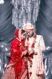 A couple stands closely together, dressed in traditional Indian wedding attire. The bride is wearing a bright red lehenga with intricate embroidery and has a veil covering her head. The groom is in a cream sherwani with a matching turban. Both are adorned with flower garlands. They appear to be in a decorative setting with draped fabric and hanging floral arrangements in the background.