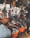 A bustling outdoor market scene with several people engaged in browsing and selling clothing items. Various garments are hanging from the stalls, and large orange tubs on the ground are filled with clothes. The market is crowded, and the atmosphere is lively with interactions among the people.