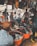 A bustling outdoor market scene with several people engaged in browsing and selling clothing items. Various garments are hanging from the stalls, and large orange tubs on the ground are filled with clothes. The market is crowded, and the atmosphere is lively with interactions among the people.