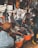 A bustling outdoor market scene with several people engaged in browsing and selling clothing items. Various garments are hanging from the stalls, and large orange tubs on the ground are filled with clothes. The market is crowded, and the atmosphere is lively with interactions among the people.