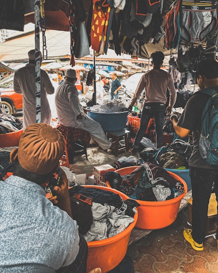 A bustling outdoor market scene with several people engaged in browsing and selling clothing items. Various garments are hanging from the stalls, and large orange tubs on the ground are filled with clothes. The market is crowded, and the atmosphere is lively with interactions among the people.