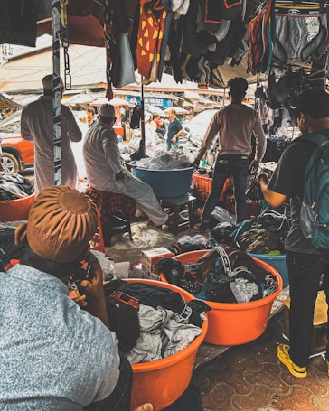 A bustling outdoor market scene with several people engaged in browsing and selling clothing items. Various garments are hanging from the stalls, and large orange tubs on the ground are filled with clothes. The market is crowded, and the atmosphere is lively with interactions among the people.