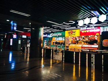 A vibrant food court area with a variety of food outlets, including familiar brands like Burger King and KFC. Bright neon signage and colorful displays attract customers, with a few people seen dining and walking. The atmosphere is lively, and the lighting is modern, highlighted by geometric ceiling lights.