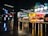 A vibrant food court area with a variety of food outlets, including familiar brands like Burger King and KFC. Bright neon signage and colorful displays attract customers, with a few people seen dining and walking. The atmosphere is lively, and the lighting is modern, highlighted by geometric ceiling lights.