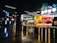 A vibrant food court area with a variety of food outlets, including familiar brands like Burger King and KFC. Bright neon signage and colorful displays attract customers, with a few people seen dining and walking. The atmosphere is lively, and the lighting is modern, highlighted by geometric ceiling lights.
