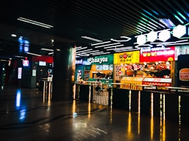 A vibrant food court area with a variety of food outlets, including familiar brands like Burger King and KFC. Bright neon signage and colorful displays attract customers, with a few people seen dining and walking. The atmosphere is lively, and the lighting is modern, highlighted by geometric ceiling lights.
