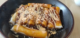 A black bowl contains rice noodle rolls drizzled with a dark sauce, cream-colored sauce, and sprinkled with sesame seeds and crushed nuts.