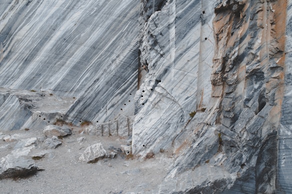 A rugged marble quarry with exposed stone faces displaying streaks of grey and white. The surface is uneven and dotted with drill holes, suggesting ongoing extraction activities. The terrain is rocky with scattered boulders and sparse vegetation.