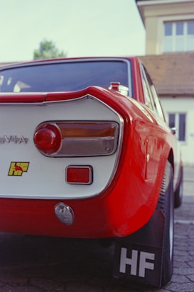 A vintage red car with distinctive tail lights and an 'HF' badge. The car is positioned on a brick-paved driveway in front of a building.
