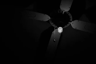 A ceiling fan with four blades and a single illuminated light bulb is suspended from the ceiling. The image is in black and white, with strong contrasts between light and dark areas, highlighting the fan's structure and the pull chain hanging from it.